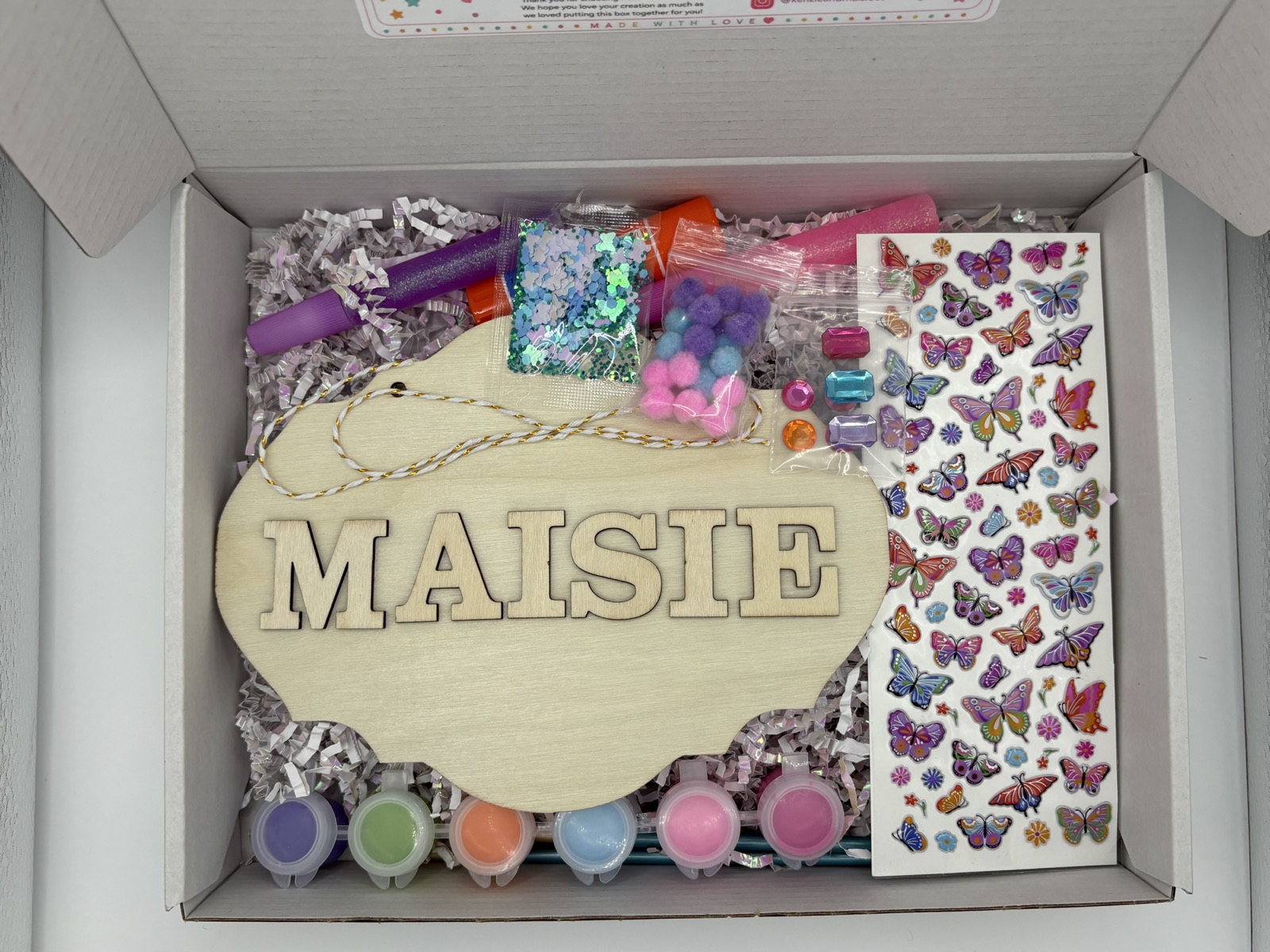 Open personalized butterfly name-sign craft kit with a wooden Maisie plaque, paints, brushes, gems, pom-poms, and stickers